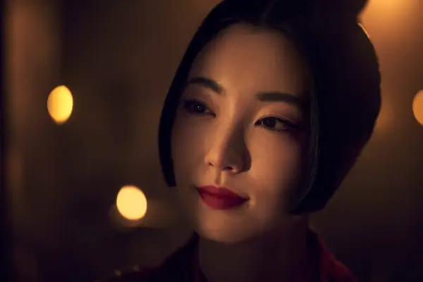 Yuka Kouri as Kiku standing in a dark room but looking towards a sliver of light, wearing red lipstick and has her hair up.