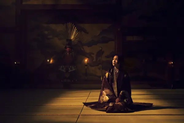 Fumi Mikado as Ochiba no Kata sitting down cross-legged in an empty room on a wooden floor and wearing a purple robe, looking up at the sky.