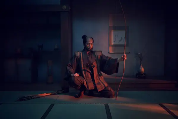 Shinnosuke Abe as Buntaro sitting in a dark room being illuminated by red light, holding a bow and arrow.