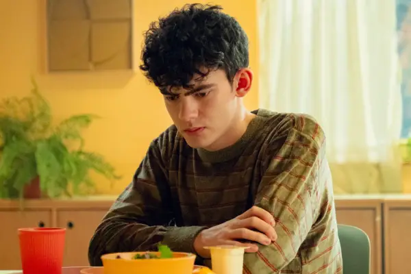 Joe Locke in Heartstopper season 3 sitting at a table and looking sad as he crosses his arms.