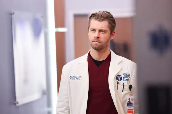 Luke Mitchell as Dr. Mitch Ripley staring off into the distance wearing red scrubs and a white lab coat.