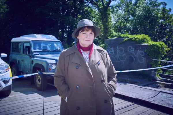 Brenda Blethyn as Vera Stanhope in Vera season 13 wearing a brown coat and hat, with a Jeep in the background