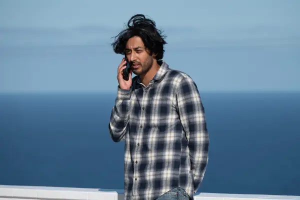 Karan Gill plays Bilal in Frauds; seen here on the phone against an ocean backdrop with wind blowing in his hair