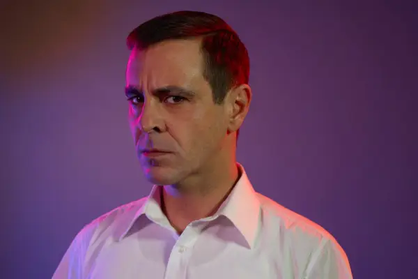 A studio photograph of Emun Elliott in character as Don Logan (from Sexy Beast). He wears a plain white shirt and stares disconcertingly down the camera lens, with a purple backdrop behind him and a purple/red light shining from above