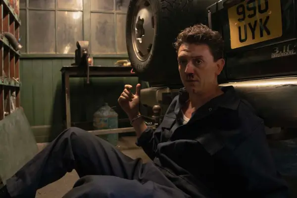 Aitch is sitting on the floor, handcuffed to the rear bumper of a car, with a bloodied nose