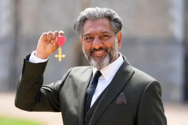 Nitin Ganatra wearing a black suit and white shirt, smiling to the camera and holding his OBE medal in his hand