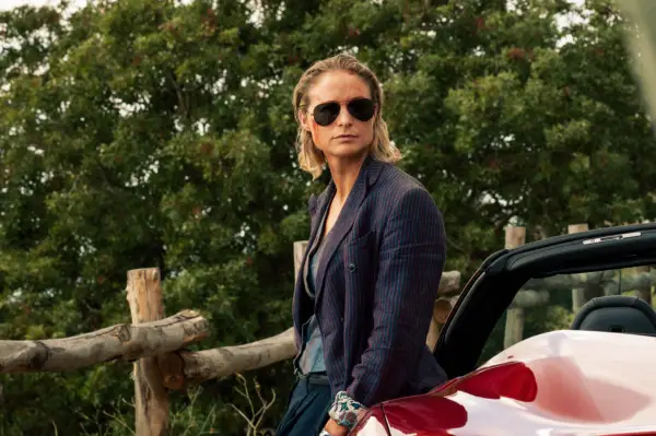 Niamh Algar as Iris in The Iris Affair, with blood on her face, wearing dark sunglasses and standing outside a car.