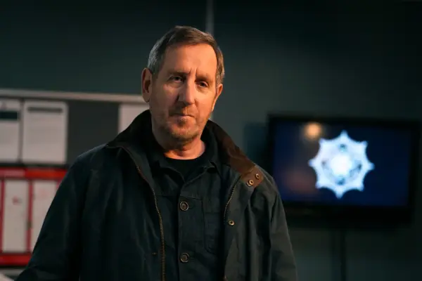 Michael Smiley as Colly in Blue Lights season 3 standing in the police station and wearing a jacket, staring at someone.