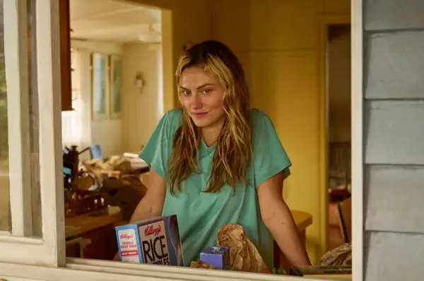 Phoebe Tonkin as Frances Bell in Boy Swallows Universe looking out of the window and leaning against the windowsill in a blue t-shirt.