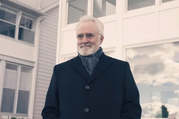 Bradley Whitford as Commander Lawrence, weraing a black coat and suit, standing outside a house, smiling