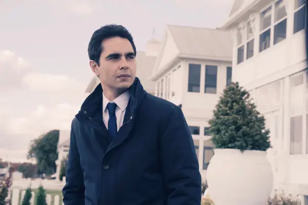 Max Minghella plays Nick, wearing a dark suit and coat, standing in a street, looking to his right