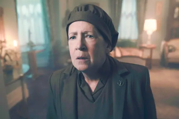 Ann Dowd plays Aunt Lydia, wearing dark clothing and a hat, with a shocked expression on her face