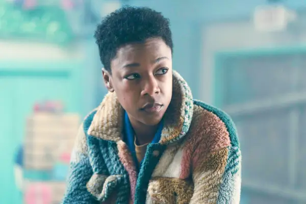 Samira Wiley plays Moira, sat down, looking to her right with a sad expression on her face, wearing a checked jacket