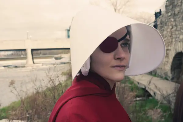 Madeline Brewer plays Janine, standing next to a river, looking at a wall, wearing a red cloak and white bonnet