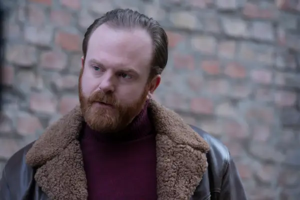 Mayhew stood outside in front of a brick wall, wearing a leather bomber jacket