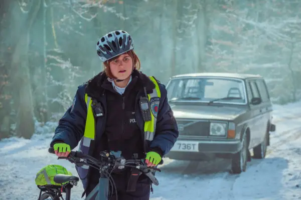 Ella Broccoleri as Ali Day pushing her bicycle along a snowy road wearing a police uniform and high vis vest in Passenger.
