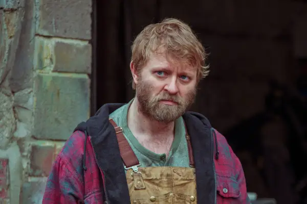 Hubert Hanowicz as Eddie Wells wearing overalls and a checkered shit looking sad in Passenger.