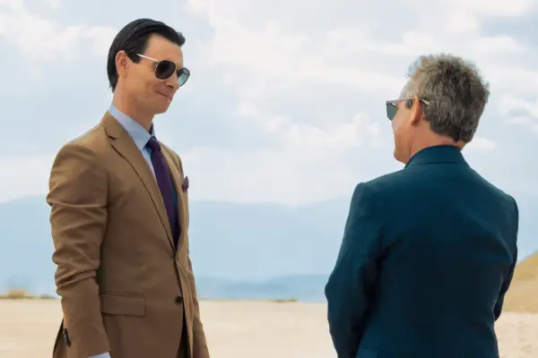 Harry Lloyd and Tom Hollander in The Iris Affair. They are both wearing suits and sunglasses and stood outside.