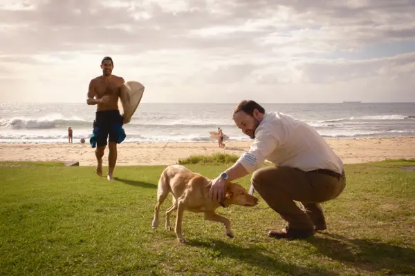 A man pets a dog near the beach while another man walks towards him
