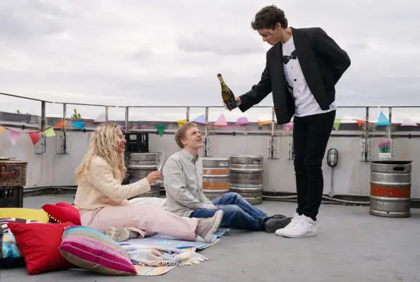 Freddie holds out a bottle of bubbly to a sitting Anna and Bobby in EastEnders