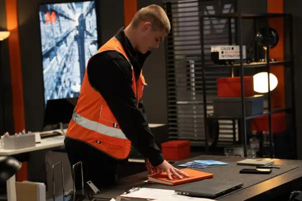 Dylan wearing a hi-vis jacket and picking up some paperwork from a desk in Emmerdale