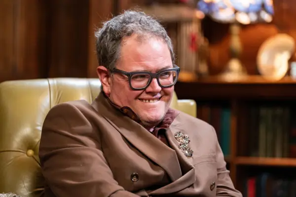 Alan Carr wearing a brown blazer, sat down in a big yellow chair smiling ahead.