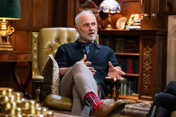Mark Bonnar wearing a black shirt and cream trousers, looking ahead.