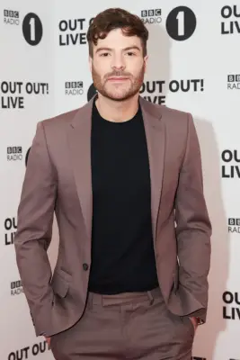 Jordan North in a black t-shirt and brown blazer with his hands in his pockets looking ahead.