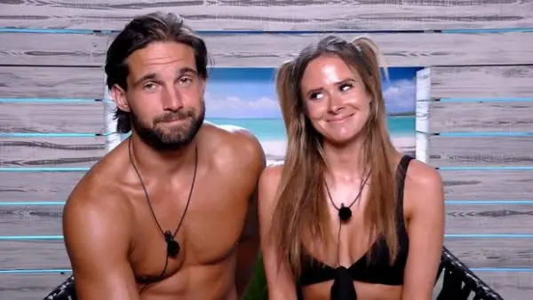 Jamie Jewitt and Camilla Thurlow sat next to each other, smiling. Jamie is topless and Camilla is wearing a black bikini and they both have black microphones around their necks.