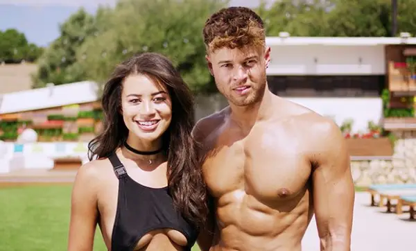 Montana Brown and Alex Beattie stood next to each other, smiling. Montana is wearing a black swimsuit while Alex is topless.
