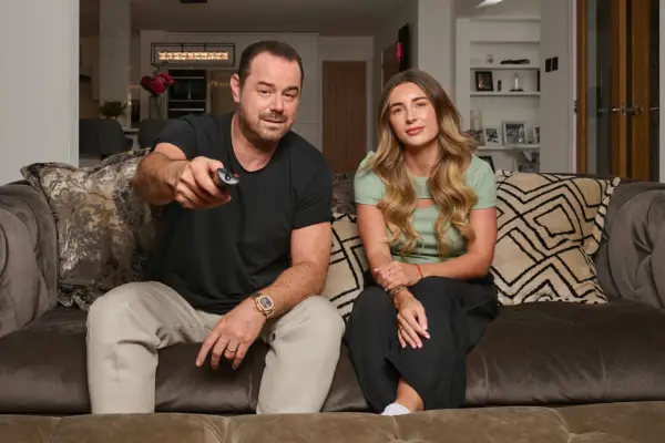Danny and Dani Dyer looking ahead at something. Danny is holding a TV remote and is pointing it forwards.