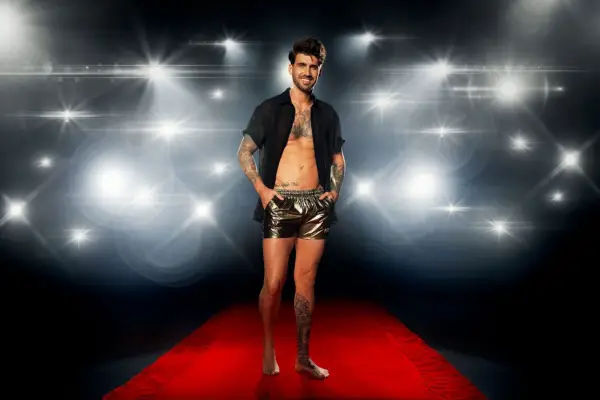 Chris Taylor on a red carpet in front of a flashing camera backdrop, wearing a black open shirt and metallic swimming shorts for Love Island: All Stars