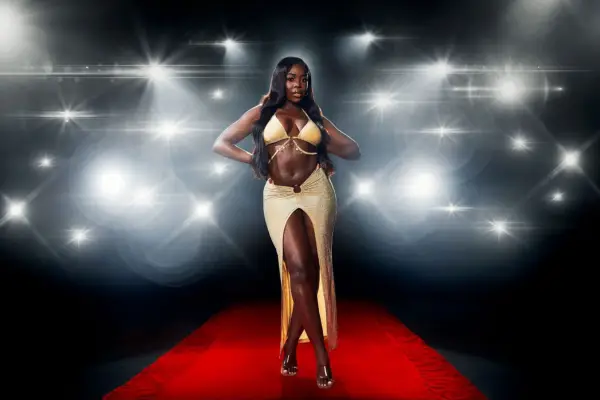 Kaz Kamwi posing on a red carpet in front of a flashing camera backdrop, while wearing a gold bra and matching skirt for Love Island: All Stars.