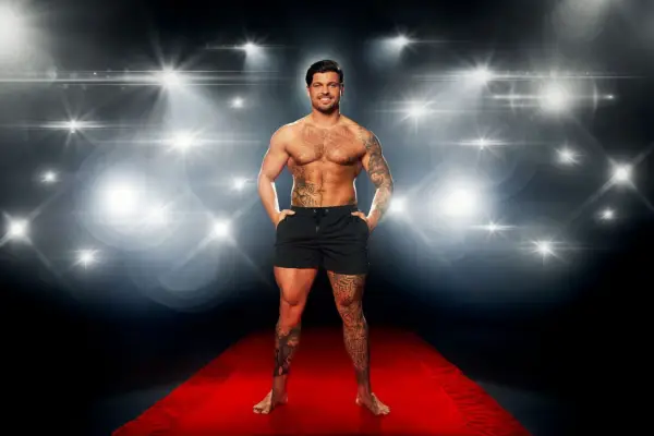 Jake Cornish posing on a red carpet in front of a flashing camera backdrop while wearing a pair of black swimming trunks.