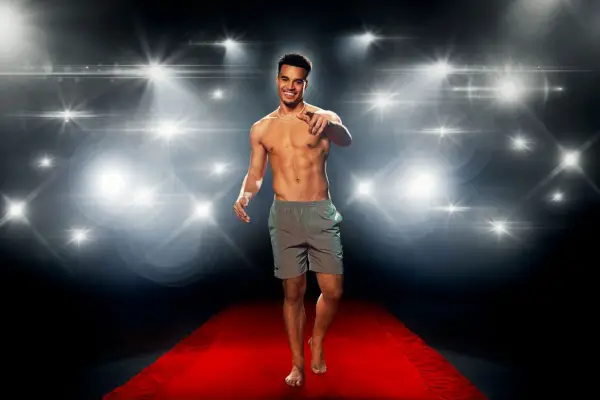 Toby Aromolaran in front of a flashing camera backdrop, wearing a pair of swimming shorts while standing on a red carpet.