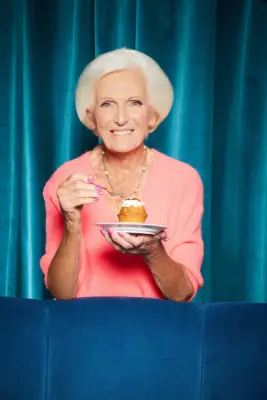 Mary Berry wearing a pink jumper holding a cake and a spoon in her hands.