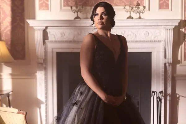 Maeve wearing a long black gown, looking to the side with her hands placed together.