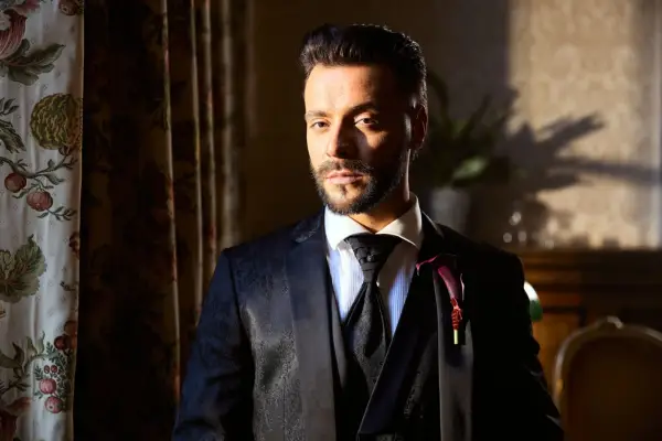 A close-up of Davide, wearing a black suit and tie looking ahead.