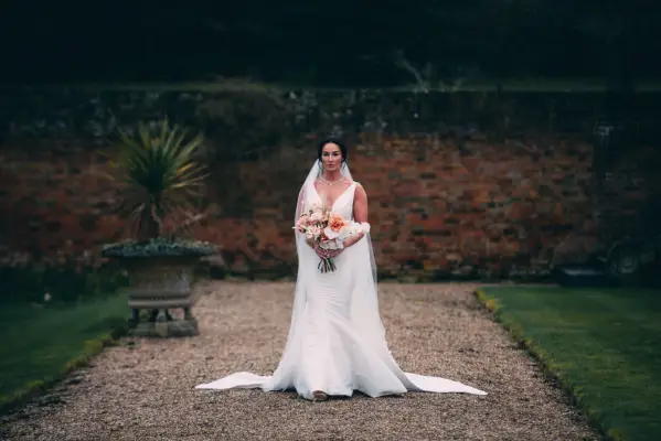 A wide shot of Rebecca, wearing a white wedding dress and veil, holding a bouquet of flowers.