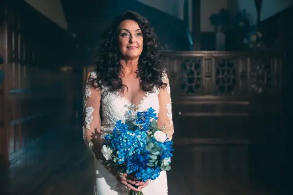 Anita wearing a white wedding dress, holding a bunch of blue flowers.