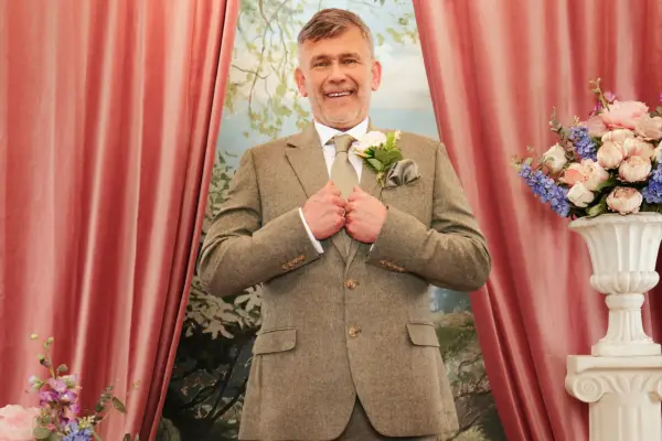 A man wearing a brown suit and tie, smiling ahead with his hands holding onto the lapels.