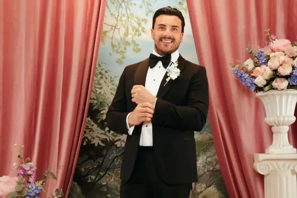 John wearing a black tuxedo, smiling ahead holding the corners of his blazer.