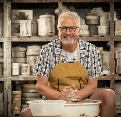 Andrew wearing a shirt and t-shirt and brown apron doing pottery