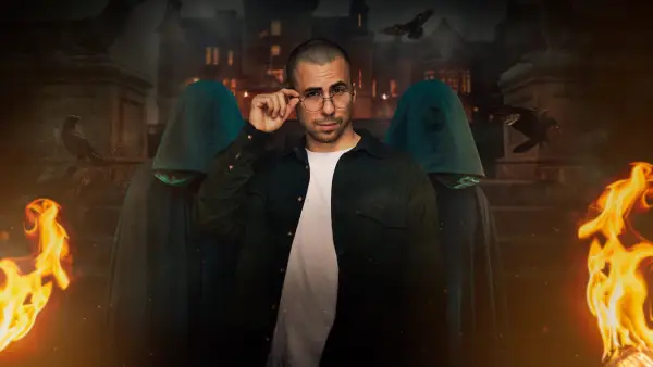 Zack for The Traitors season 2. He is wearing a white t shirt and a black shirt. He is holding onto his glasses, looking curious.