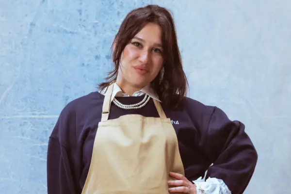 Nataliia smiling ahead in front of a light blue background. She is wearing a black jumper with a white shirt with a brown apron over it.