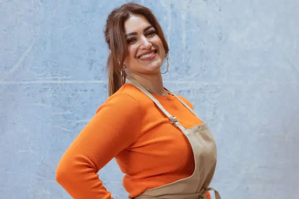 Nadia wearing an orange top and a light brown apron, smiling ahead. Her body is turned to the side with one hand on her hip.