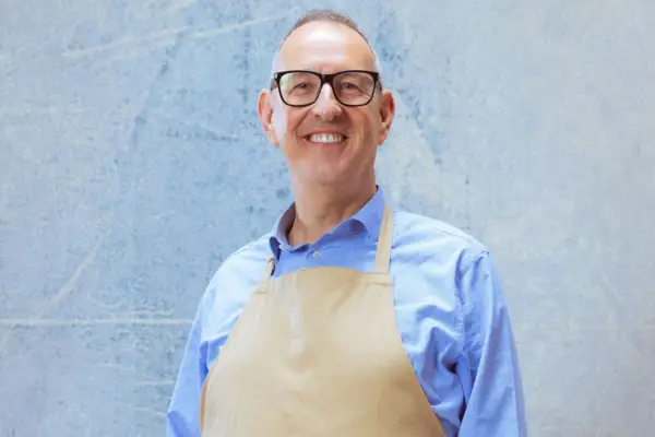 Leighton smiling ahead, wearing a blue shirt and a light brown apron.