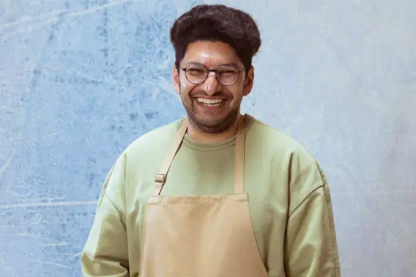 Hassan smiling ahead posing in front of a light blue background. She is wearing a light green jumper with a brown apron.