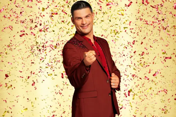 Aljaž Škorjanec wearing a dark red blazer and shirt, pointing ahead of him.