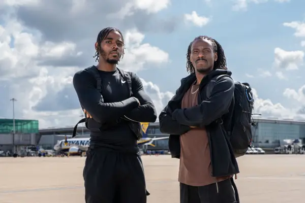 Two men stand together with their arms crossed on the runway of an airport and look into the camera.